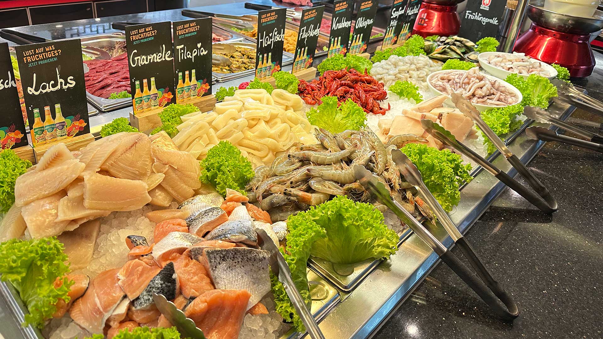 Selbstbedienungstheke mit frischem Fisch, Garnelen, Tintenfisch und Gemüse umgeben von Salatblättern.