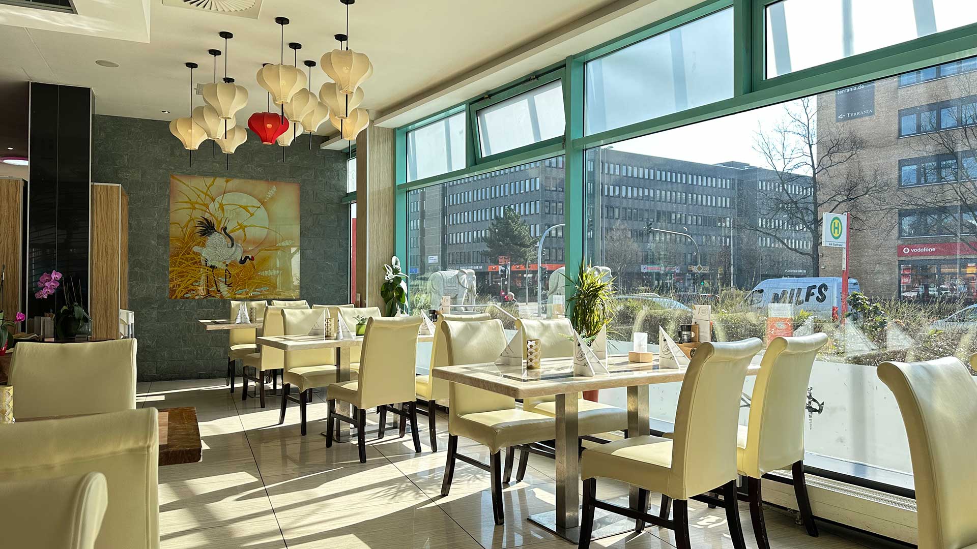 Ein modernes Café mit weißen Stühlen, großer Fensterfront und dekorativen Lampen.